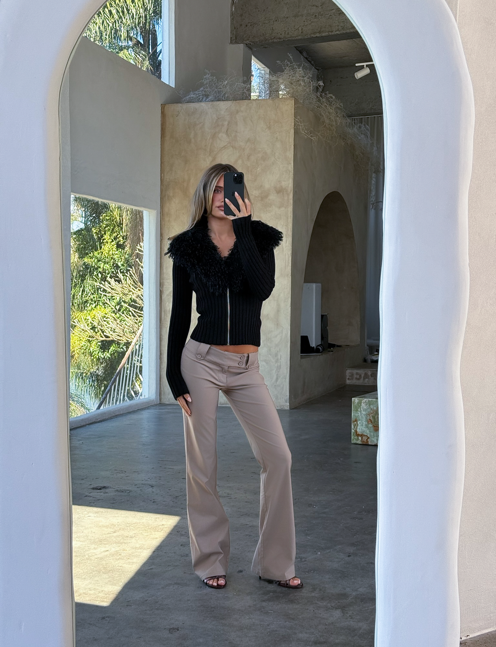 A person takes a selfie in front of a large arched mirror, wearing the TIGERMIST BRISA JUMPER - BLACK with a faux fur collar and beige pants, surrounded by minimalist decor and natural light from big windows.