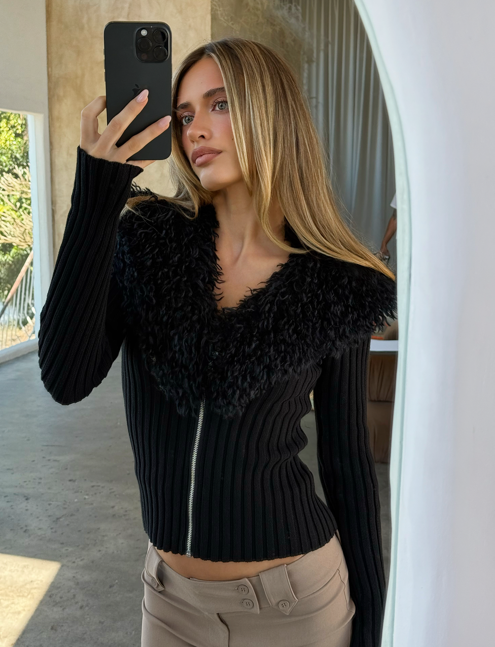 A woman with long blonde hair takes an indoor mirror selfie in sunlight, wearing the TIGERMIST BRISA JUMPER - BLACK, a knit ribbed cardigan with a faux fur collar, paired with beige pants in a minimalist setting.