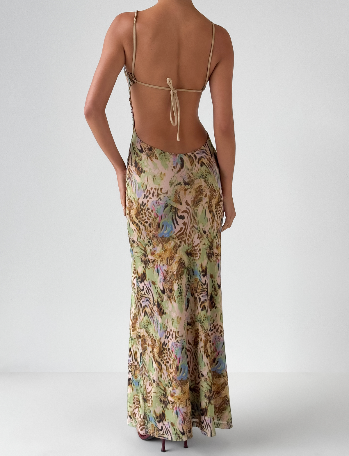 A person stands facing away, wearing the TIGERMIST ELYSIUM MAXI DRESS - ANIMAL SWIRL, a backless, spaghetti-strap, floor-length bodycon dress with a colorful animal swirl print. The background is plain white.