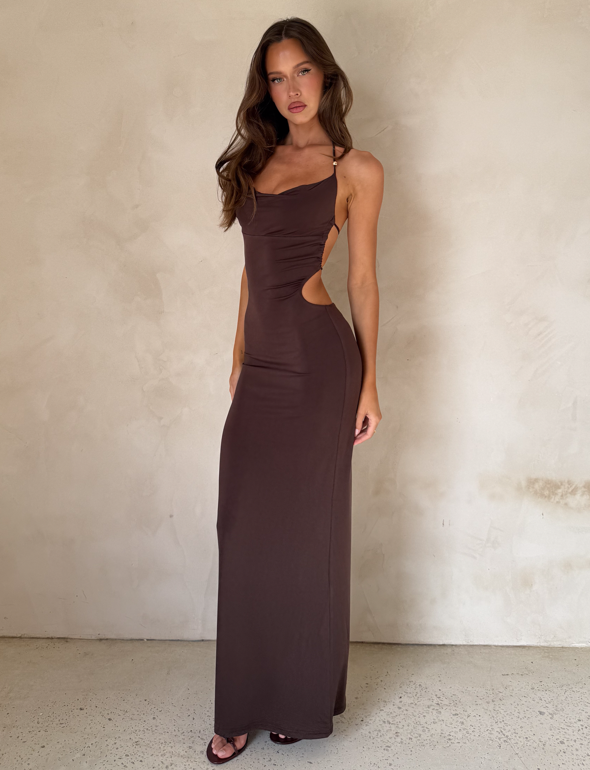 A woman with long, wavy hair and a neutral expression stands against a light wall, wearing the TIGERMIST SURIYA MAXI DRESS in chocolate brown with side cutouts, cowl neck, halter straps, gold waist chain, and black heeled sandals.