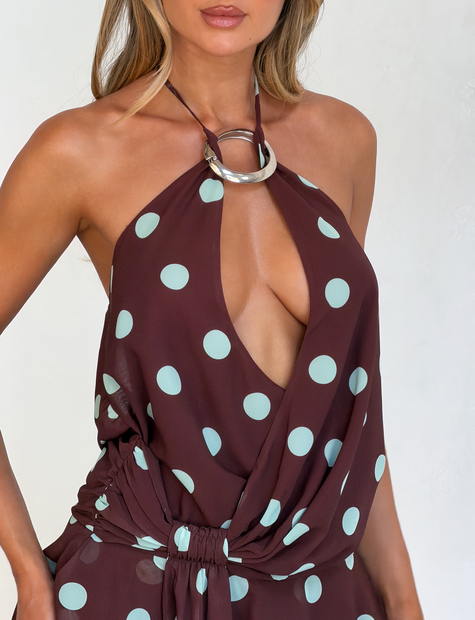 A woman wears the TIGERMIST INDIGO MINI DRESS 2.0 - CHOCOLATE BLUE DOT, a sleeveless brown halter mini with large blue polka dots, deep v-neck, gathered waist, and circular metal ring at the neckline. Her face is partially out of frame.