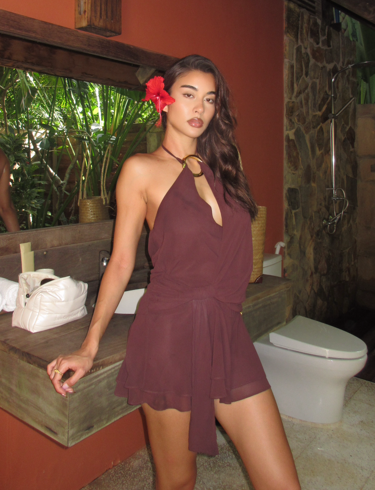 A woman wearing the TIGERMIST INDIGO MINI DRESS 2.0 in chocolate, with a self-tie waist, poses by a rustic sink and mirror in a bathroom featuring stone walls and tropical plants reflected in the glass.