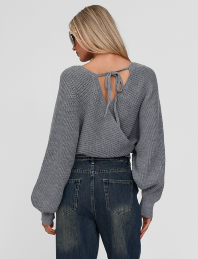 A woman with long blonde hair and sunglasses wears the TIGERMIST RAVEN JUMPER - GREY, featuring a crisscross back, tie detail, and bell sleeves, paired with high-waisted blue jeans. She faces away from the camera.
