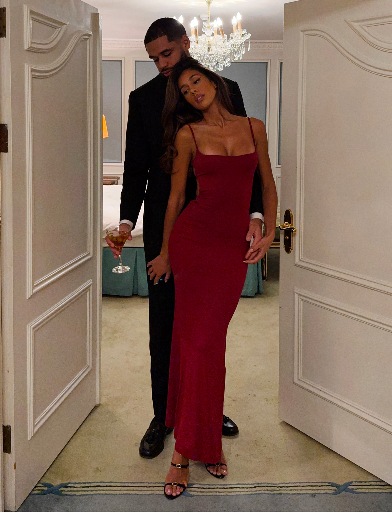 A man in a black suit embraces a woman wearing the TIGERMIST MINERVA MAXI DRESS - RED : BERRY RED; she poses gracefully under the chandelier, highlighting the dress's elegant bodycon silhouette.