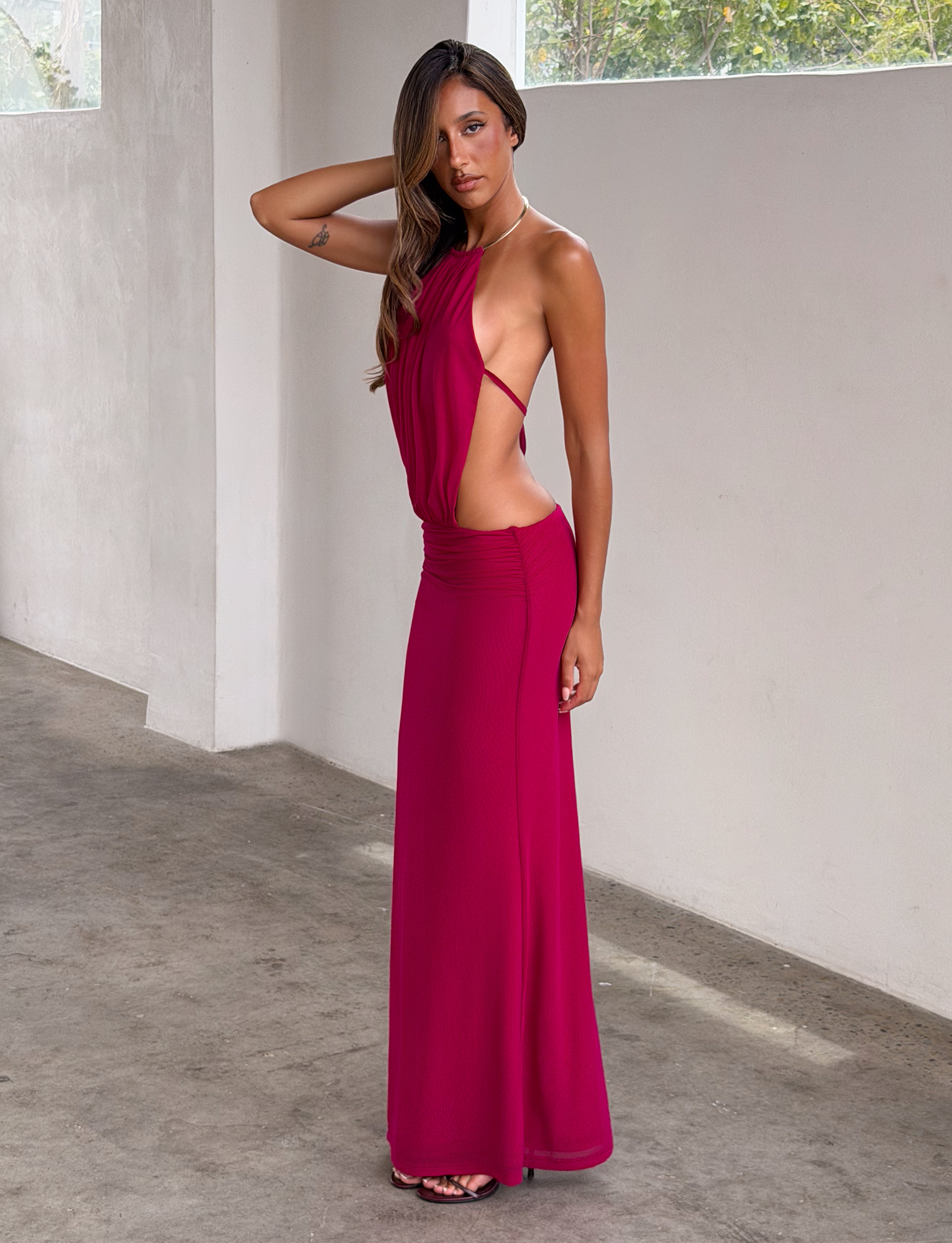A woman stands indoors on a concrete floor, wearing the TIGERMIST EVANNA MAXI DRESS in berry with side cutouts and black sandals. She has long hair, a tattoo on her upper arm, and poses with one hand touching her head.