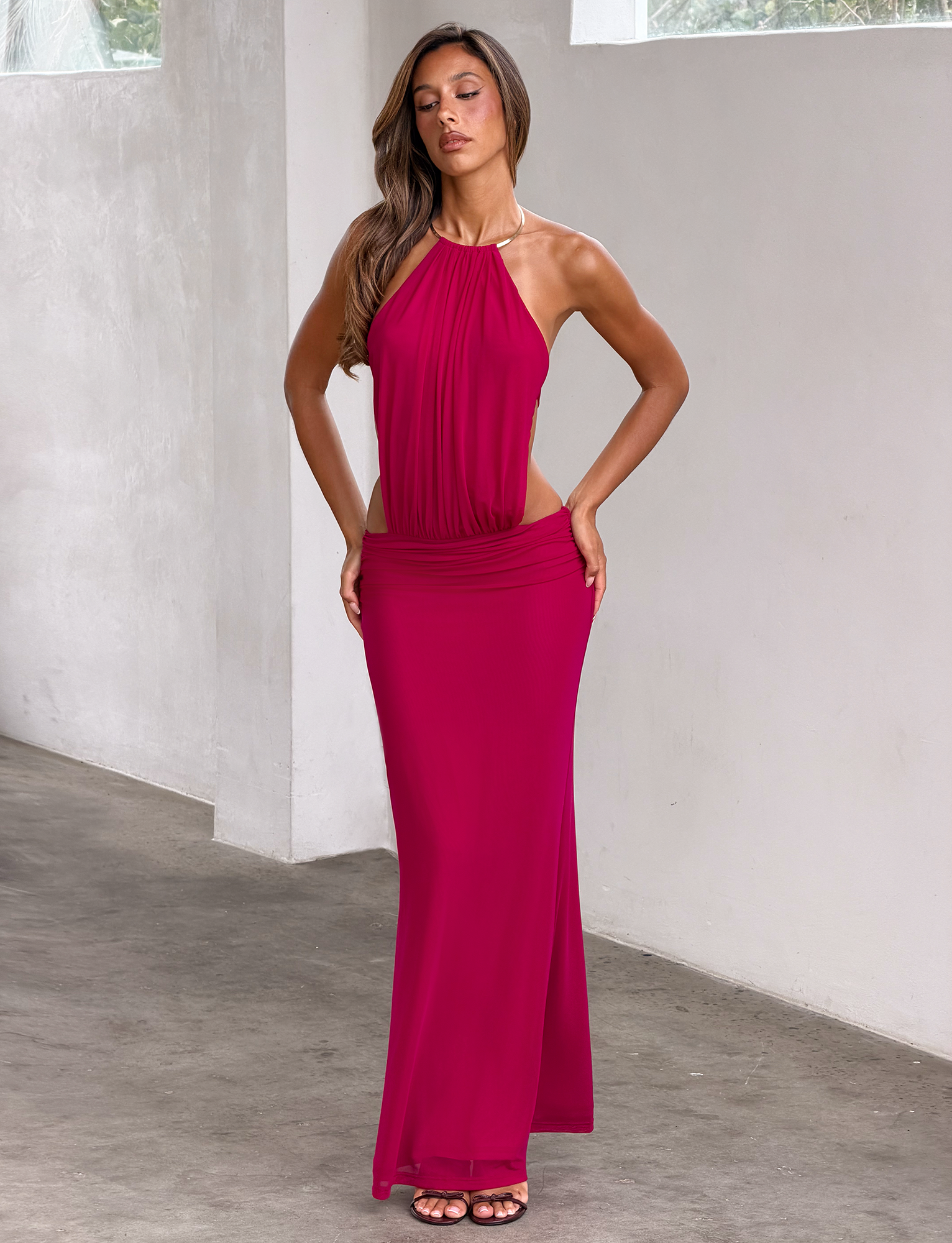 A woman with long brown hair poses indoors against a white wall, wearing the TIGERMIST EVANNA MAXI DRESS in berry—a sleeveless mesh dress with a halter neckline.