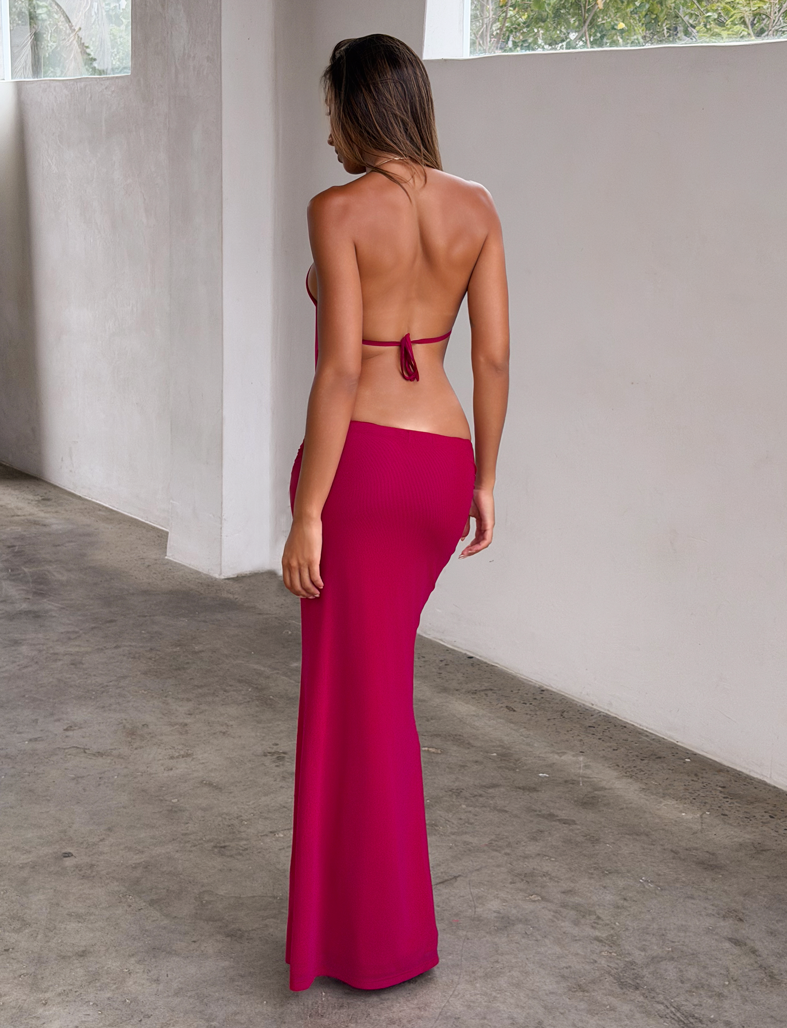 A woman with long hair stands indoors, facing away, wearing the TIGERMIST EVANNA MAXI DRESS in Berry. The open-back, halter-neck dress has a gold-trim neckline. The setting has white walls, concrete floors, and sunlit large windows.