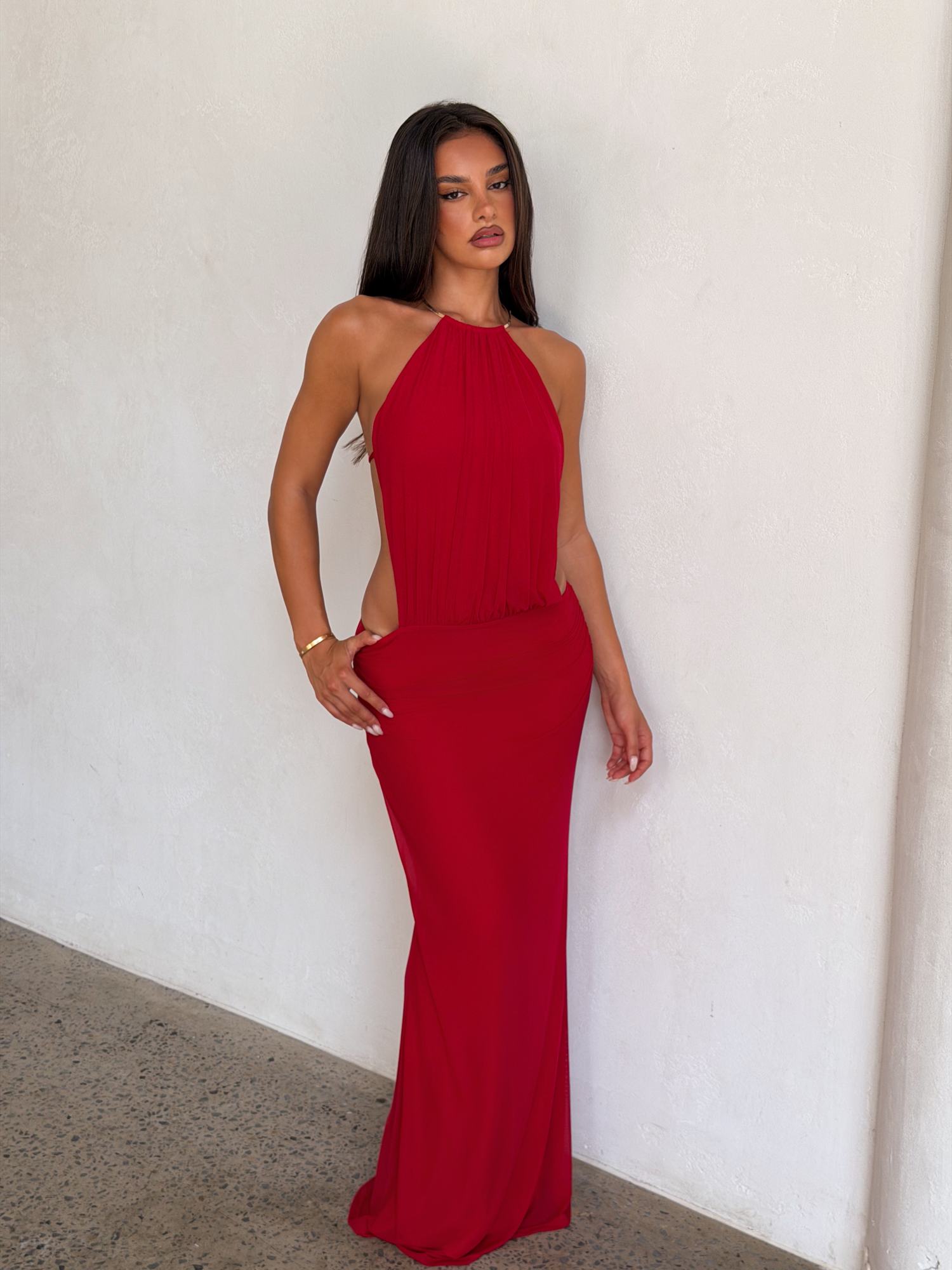 A woman with long dark hair stands against a light wall, wearing the TIGERMIST EVANNA MAXI DRESS in red—a sleeveless, high-neck, floor-length dress. She has a neutral expression and wears minimal jewelry, including a bracelet on her right wrist.