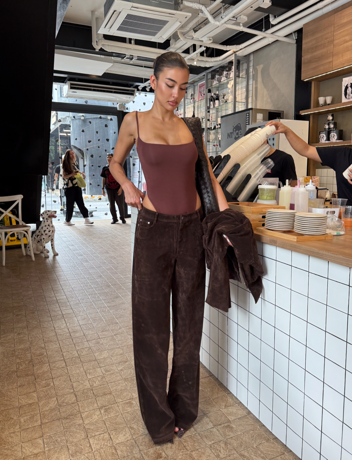 A woman in TIGERMIST's CAMELLIA PANT - CHOCOLATE SUEDE stands by a tiled café counter, holding a matching jacket. Her hair is tied back as she looks down; shelves, baristas, and people by large windows fill the background.