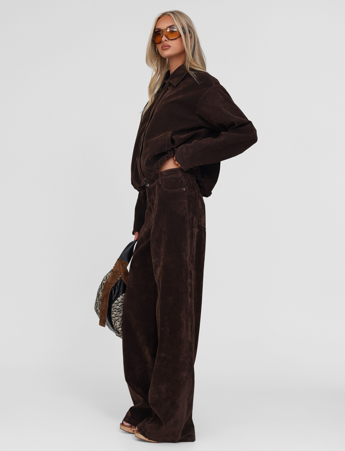 A woman stands in profile against a white background wearing TIGERMIST's CAMELLIA PANT in chocolate suede with a matching long-sleeve shirt, holding a patterned bag and sunglasses.