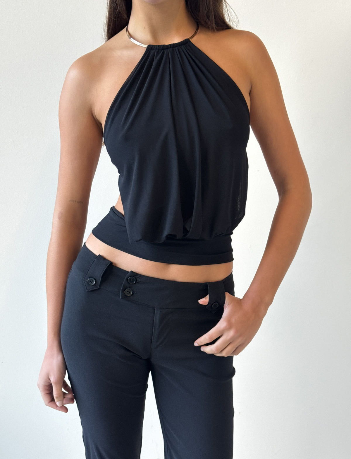 A person wears the TIGERMIST EVANNA TOP in black—a sleeveless halter style with a gathered bodice—paired with black pants featuring button details, standing against a plain light background with their right hand on their hip.