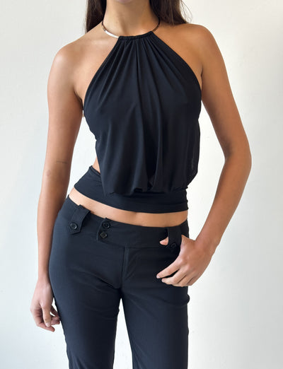 A person wears the TIGERMIST EVANNA TOP in black—a sleeveless halter style with a gathered bodice—paired with black pants featuring button details, standing against a plain light background with their right hand on their hip.