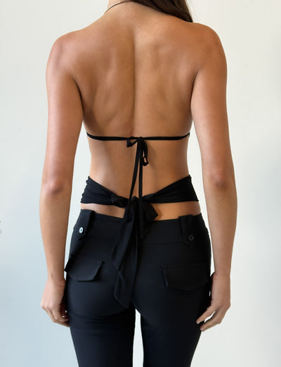 A woman is seen from the back wearing the TIGERMIST EVANNA TOP - BLACK with thin straps tied at the mid-back and waist, gathered bodice detail, paired with black pants featuring flap pockets against a plain light background.
