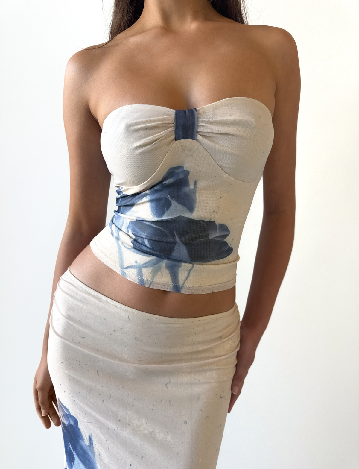 A woman wears the TIGERMIST JANIS TOP - INDIGO FLORAL, a strapless tube top in cream with abstract blue rose patterns, paired with a matching skirt, standing against a plain white background.