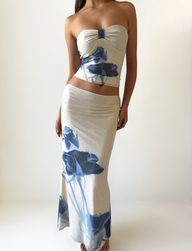 A woman models the TIGERMIST JANIS MAXI SKIRT in Indigo Floral, a beige skirt with a fitted waist and long silhouette featuring bold blue florals, standing against a plain white background.