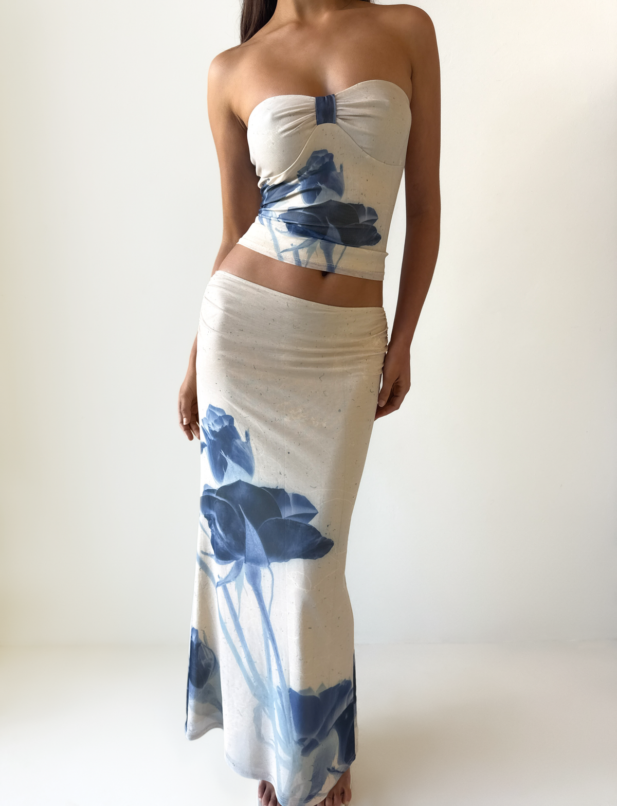 A woman models the TIGERMIST JANIS TOP - INDIGO FLORAL, a strapless tube top with blue rose print, paired with a matching long skirt in a cream two-piece set, standing against a plain white background.