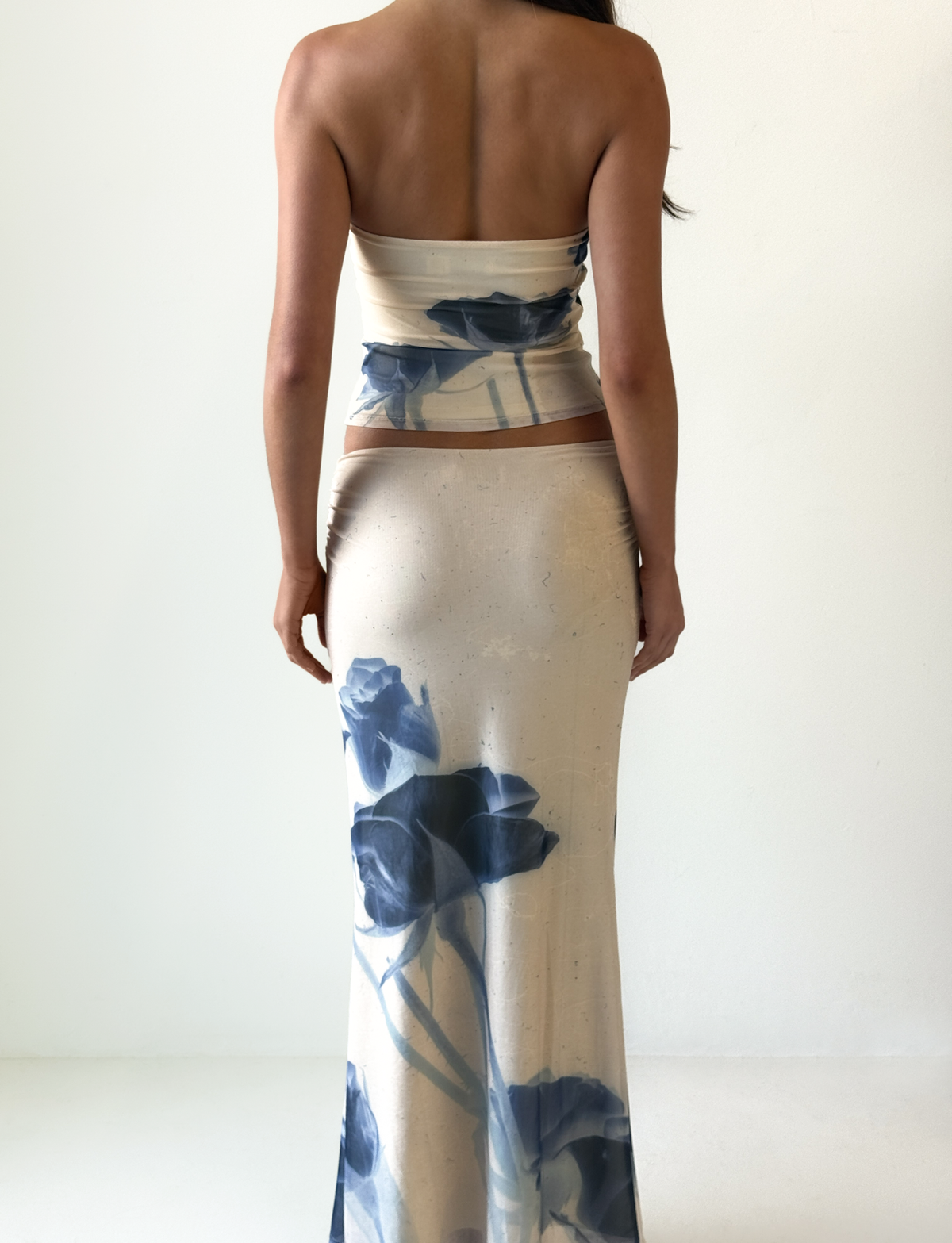 A woman stands with her back to the camera, wearing the TIGERMIST JANIS MAXI SKIRT in indigo floral. The skirt is form-fitting and features a bold blue floral pattern. The background is plain white.