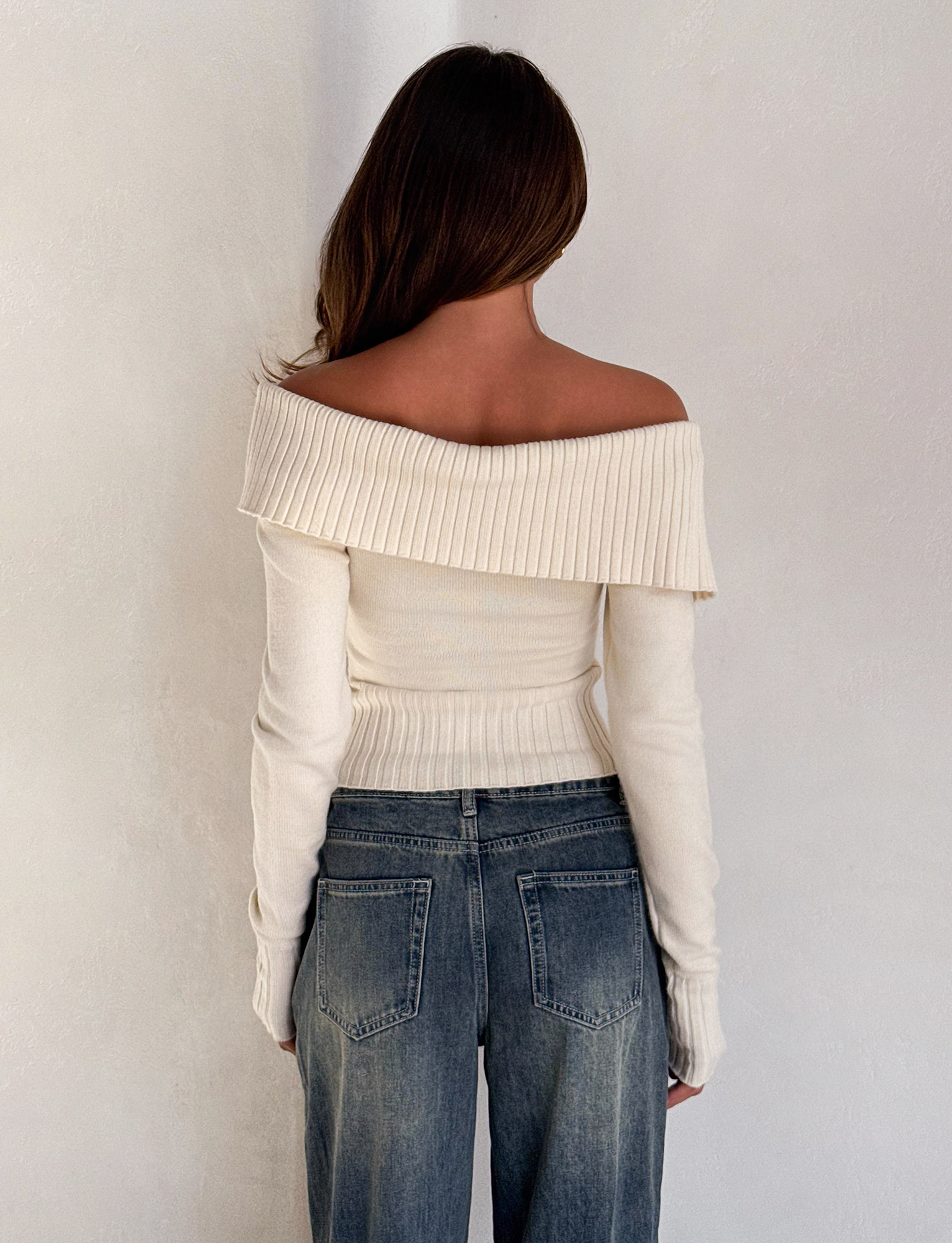 A woman with long brown hair stands facing away against a plain white wall, wearing the TIGERMIST KIERA JUMPER in cream and blue jeans.