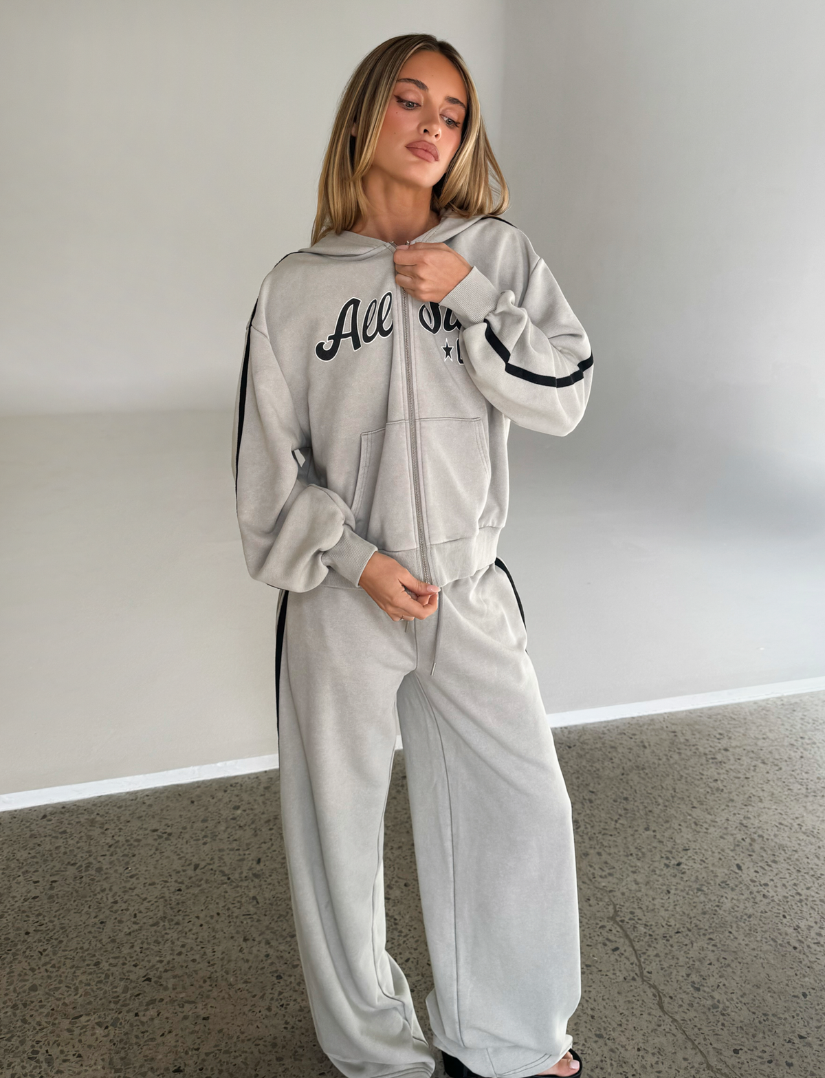 A woman stands indoors on a concrete floor wearing the TIGERMIST PAYTON HOODIE in stone with a matching tracksuit, looking slightly to the side and touching her collar.