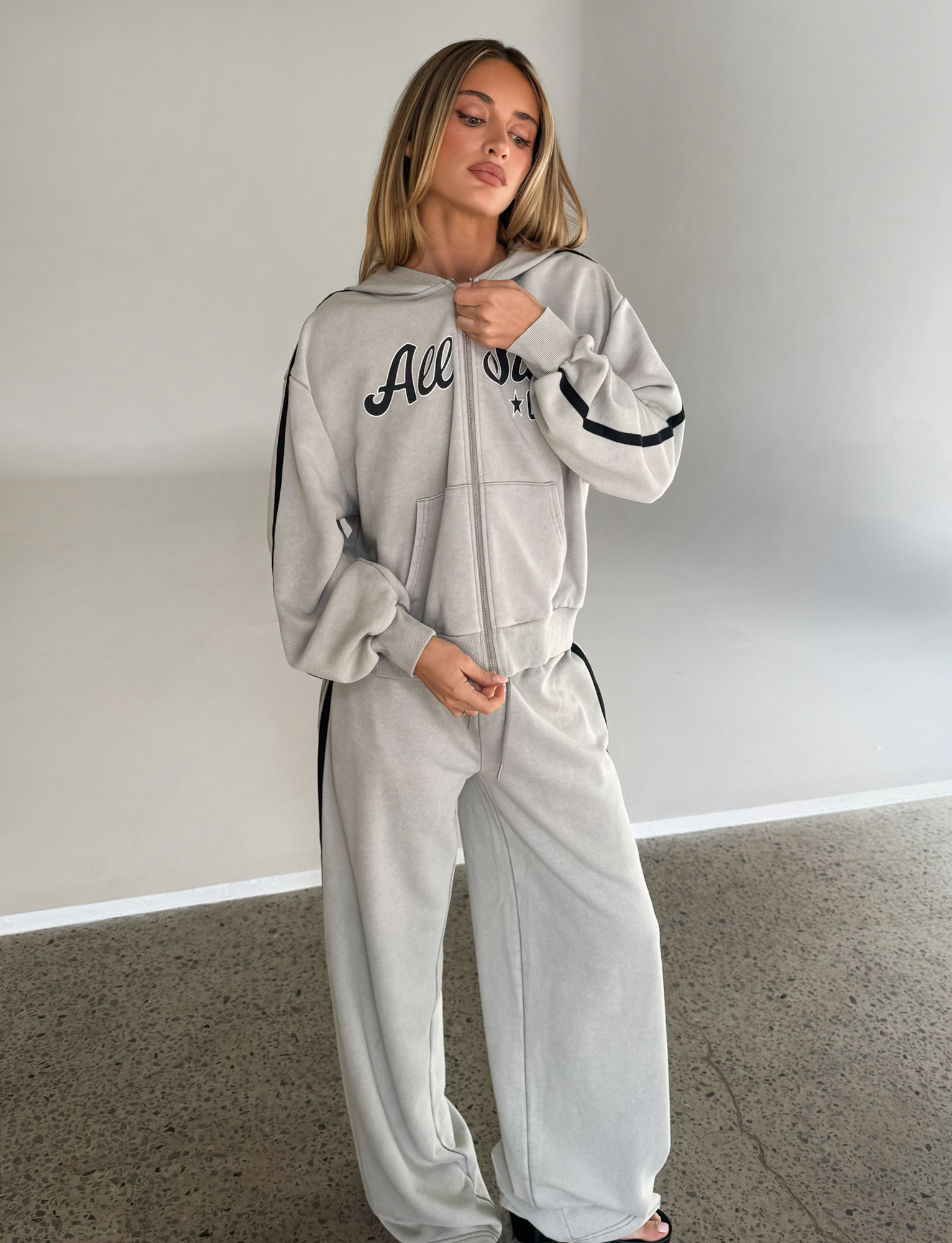 A woman stands indoors on a concrete floor wearing the TIGERMIST PAYTON HOODIE in stone with a matching tracksuit, looking slightly to the side and touching her collar.