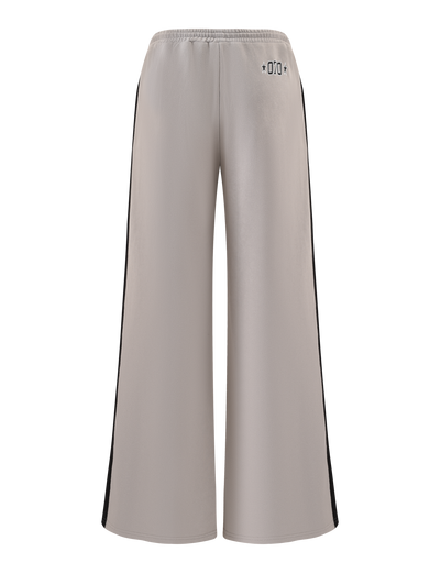 The PAYTON TRACKPANT - STONE by TIGERMIST features a wide-leg fit, taupe cotton fabric, elastic waistband, black side panels, and small black symbols near the back waist. The sporty pants are shown against a white background.