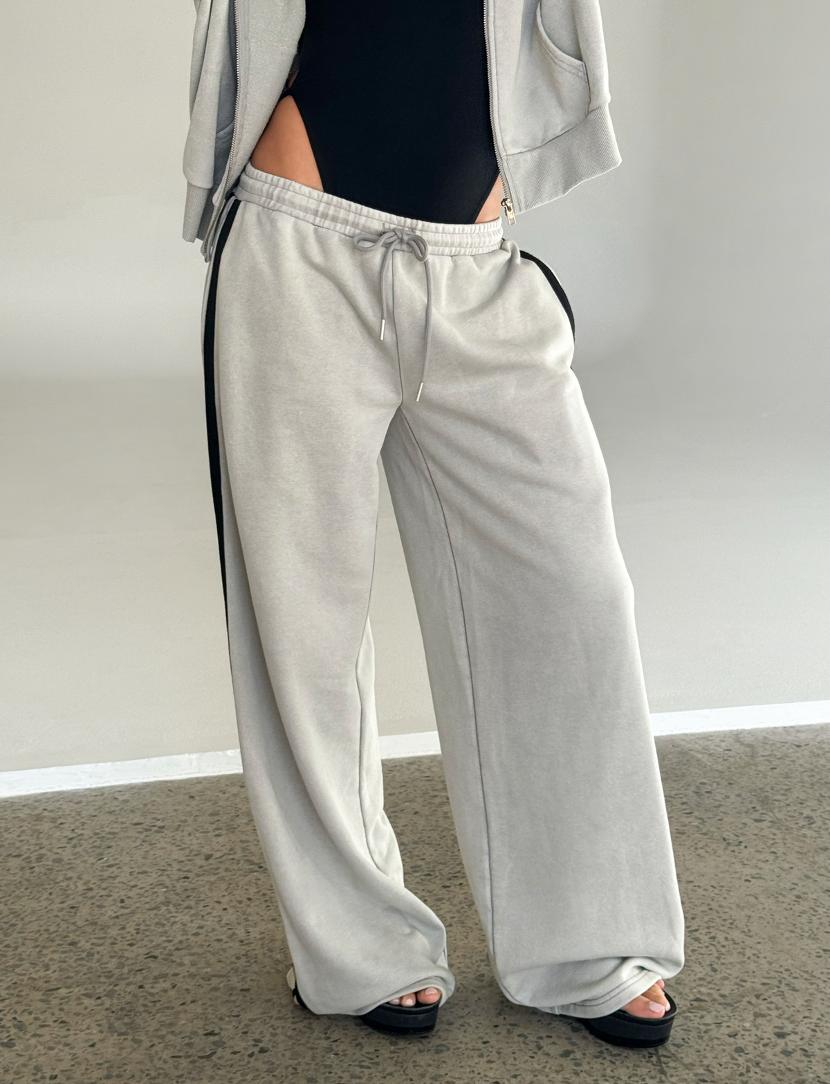A person wearing TIGERMIST PAYTON TRACKPANT in Stone, featuring a light gray color and drawstring waist, paired with a black top and platform sandals stands on a speckled floor. Only the lower body and part of the torso are shown.