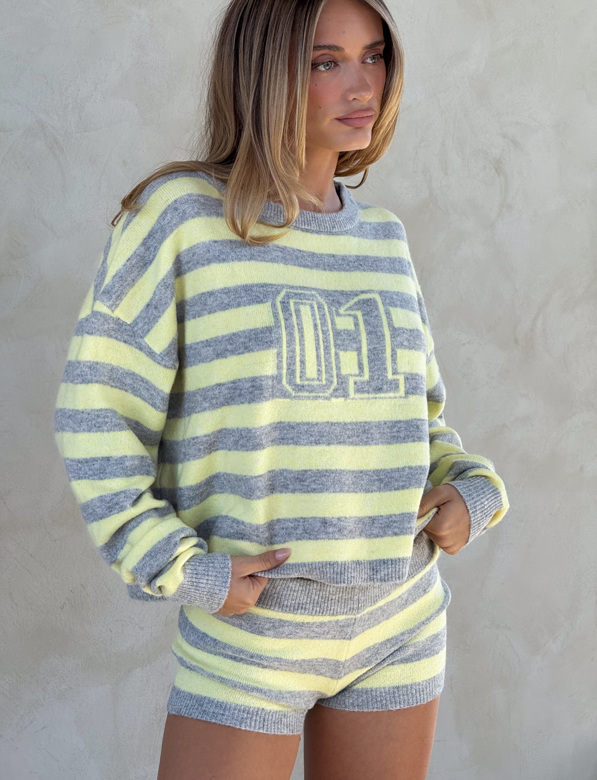 A woman wears the KOA JUMPER in yellow and grey stripes by TIGERMIST, styled oversized with a sporty "01" print, paired with matching shorts as she stands against a light-colored textured wall, hands in pockets.