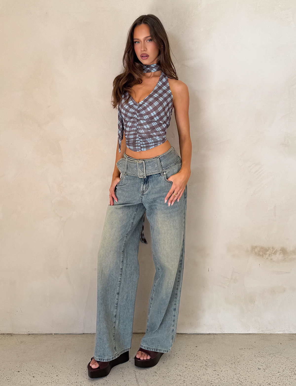 A woman poses against a textured light wall wearing TIGERMIST's REEF JEAN - LIGHTWASH, styled with a blue plaid sleeveless top, black platform sandals, a belt, and a matching neck scarf.