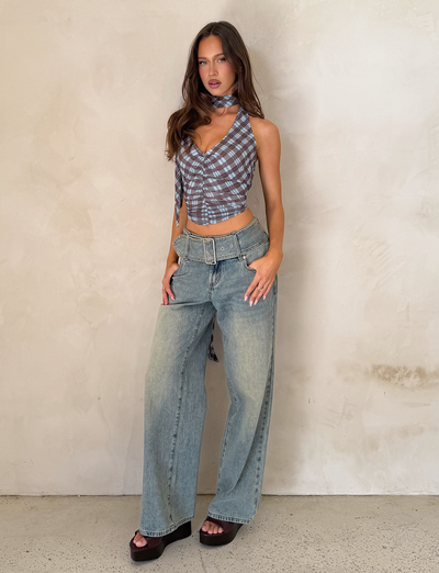 A woman poses against a textured light wall wearing TIGERMIST's REEF JEAN - LIGHTWASH, styled with a blue plaid sleeveless top, black platform sandals, a belt, and a matching neck scarf.