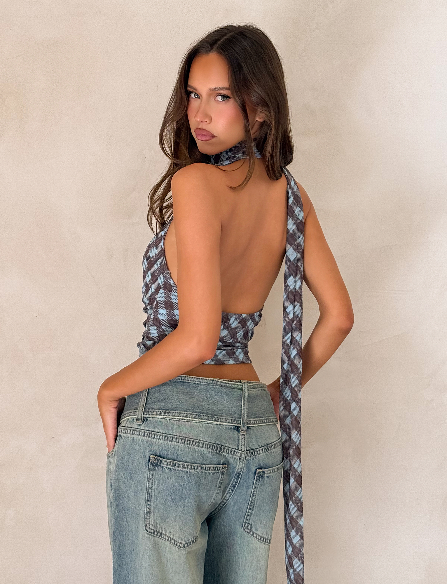 A woman with long brown hair poses with her back turned, wearing the TIGERMIST NOBLE TOP - BLUE CHECK styled as a halter neck scarf and light blue jeans against a beige textured wall.