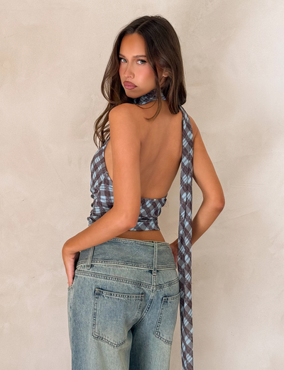 A woman with long brown hair poses with her back turned, wearing the TIGERMIST NOBLE TOP - BLUE CHECK styled as a halter neck scarf and light blue jeans against a beige textured wall.