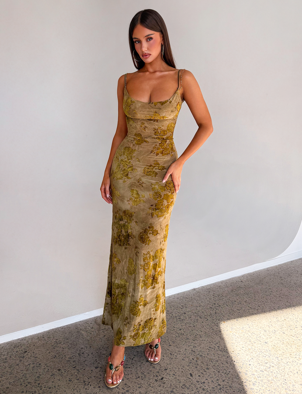 A woman poses gracefully against a plain wall wearing the TIGERMIST SPARROW MAXI DRESS in Green Burnout, a long, fitted floral piece with sheer straps, styled with open-toe sandals. Her long hair flows as she poses with one hand by her side and the other on her hip.