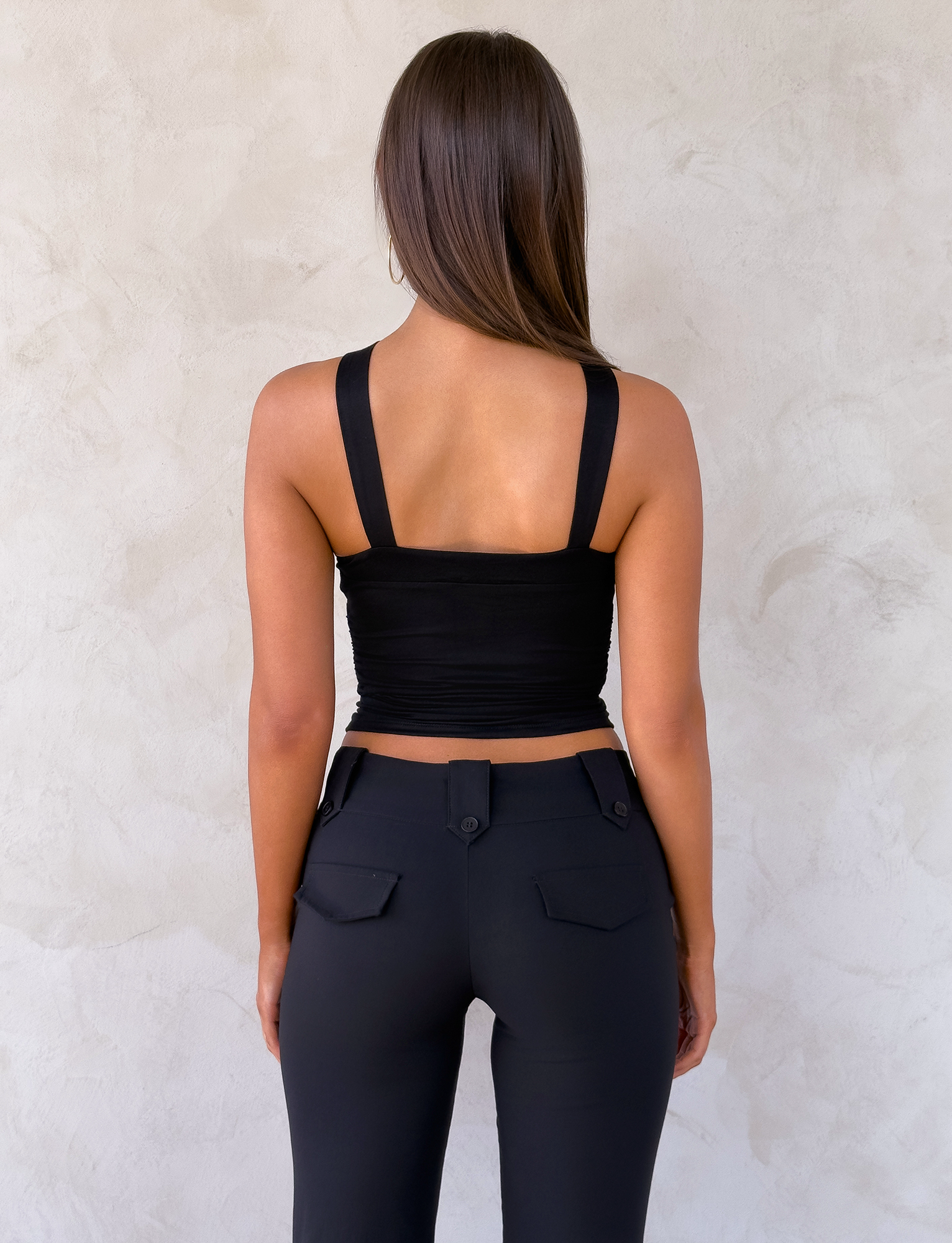 A woman with long brown hair stands facing a textured light wall, wearing TIGERMIST's FINCH TOP in black and black flap-pocket pants. Her arms are by her sides.