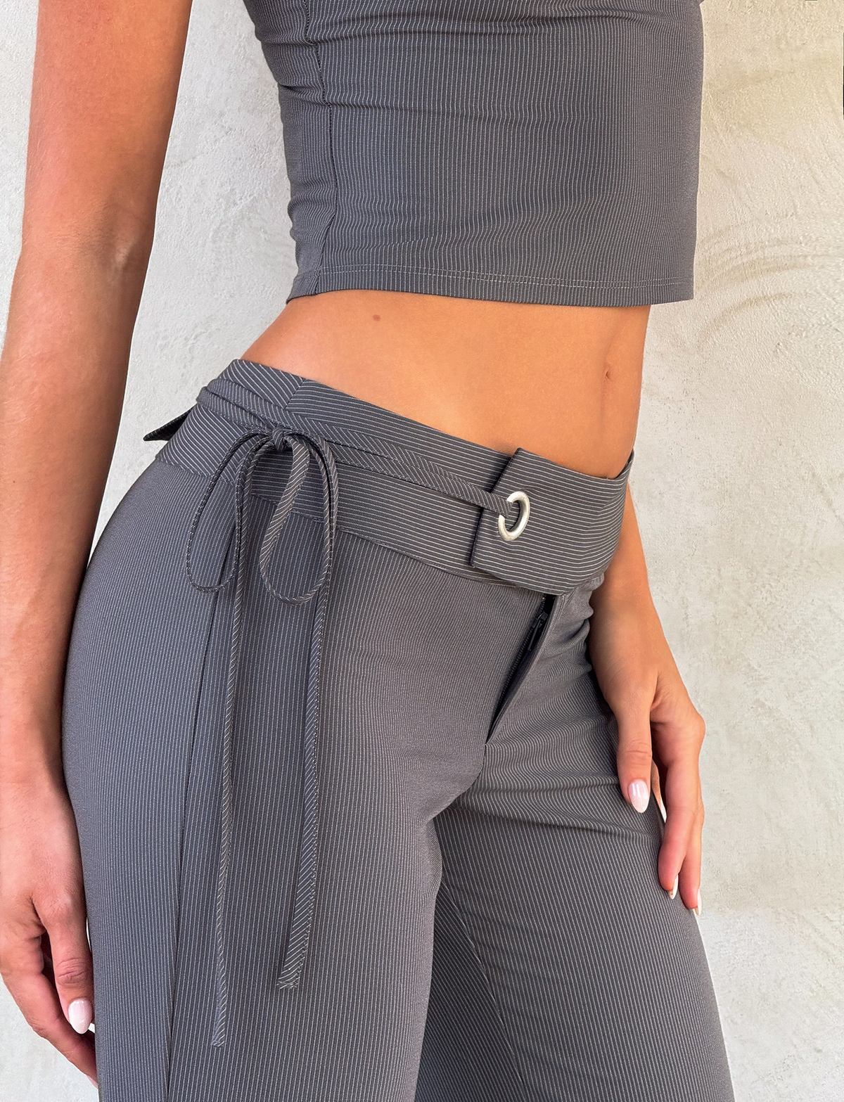 A person wears the TIGERMIST EMORY PANT in grey pinstripe with a tie belt, paired with a gray ribbed sleeveless crop top, standing against a light textured wall.