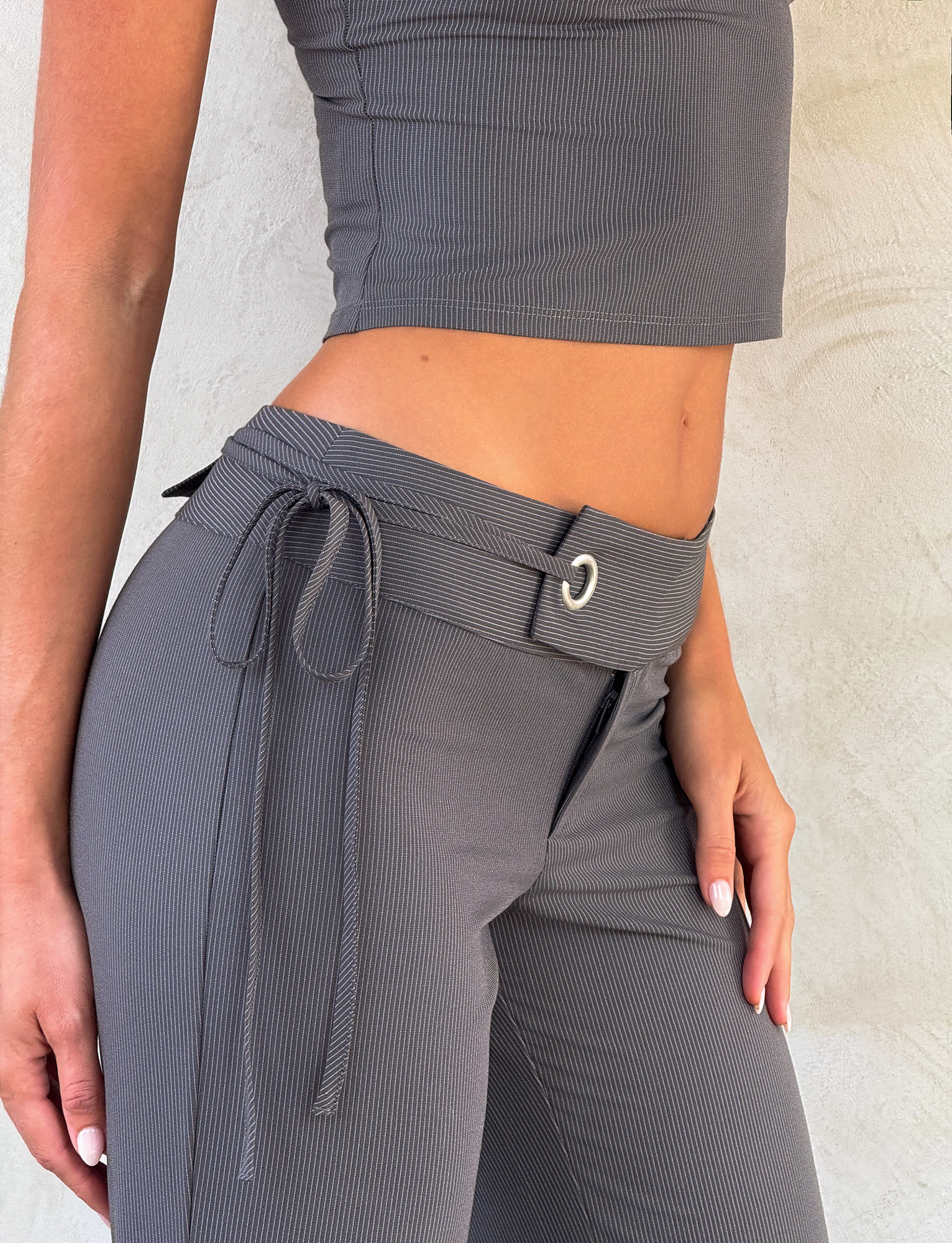 A person wears the TIGERMIST EMORY PANT in grey pinstripe with a tie belt, paired with a gray ribbed sleeveless crop top, standing against a light textured wall.