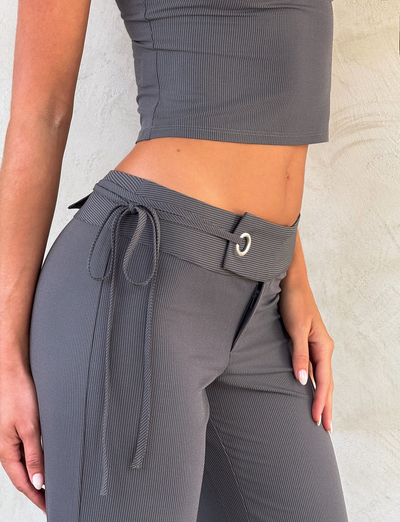 A person wears the TIGERMIST EMORY PANT in grey pinstripe with a tie belt, paired with a gray ribbed sleeveless crop top, standing against a light textured wall.
