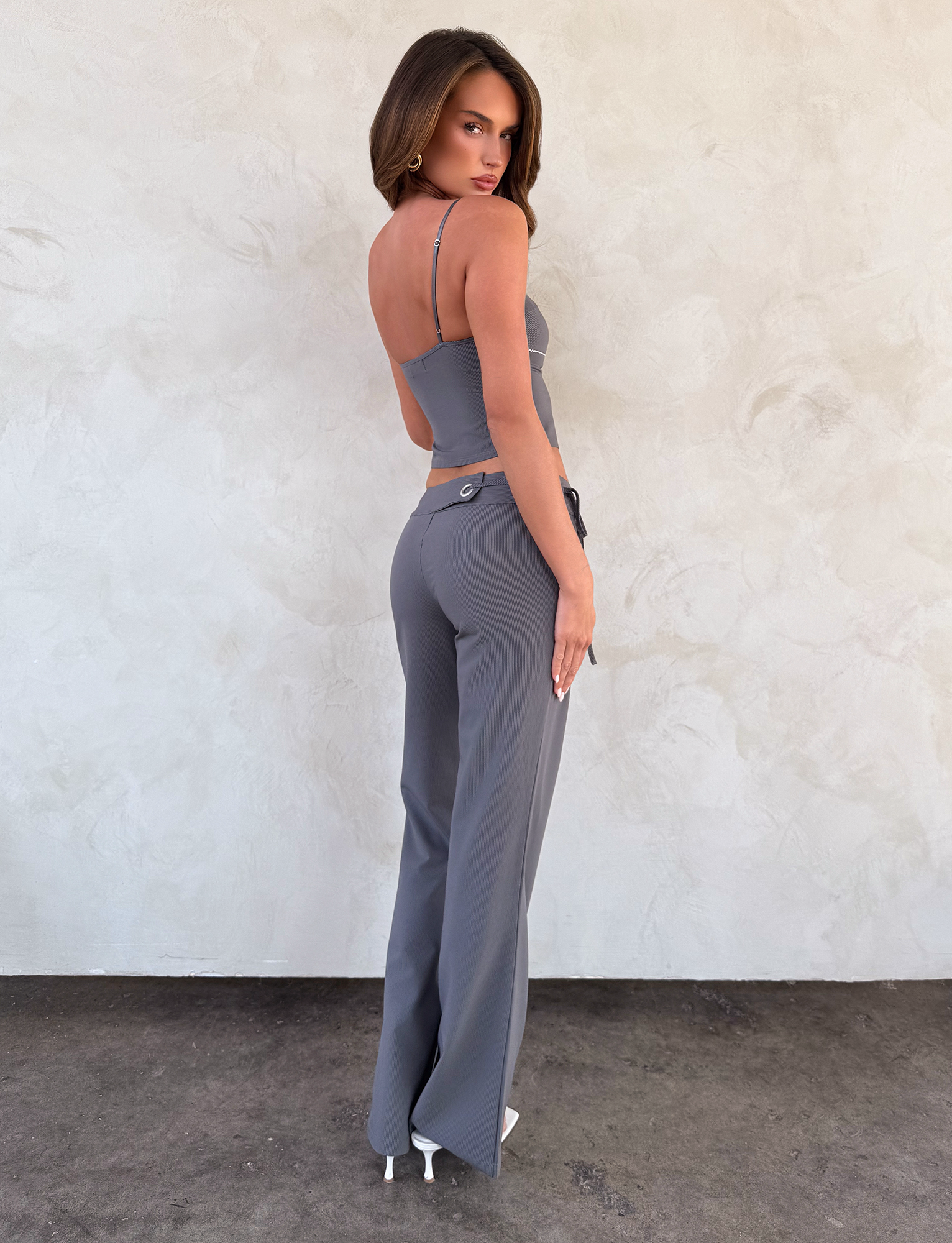 A woman wears the TIGERMIST EMORY PANT in grey pinstripe—a low rise fit—paired with a matching two-piece outfit and white heels, standing mostly with her back to the camera and glancing over her shoulder against a light textured wall.