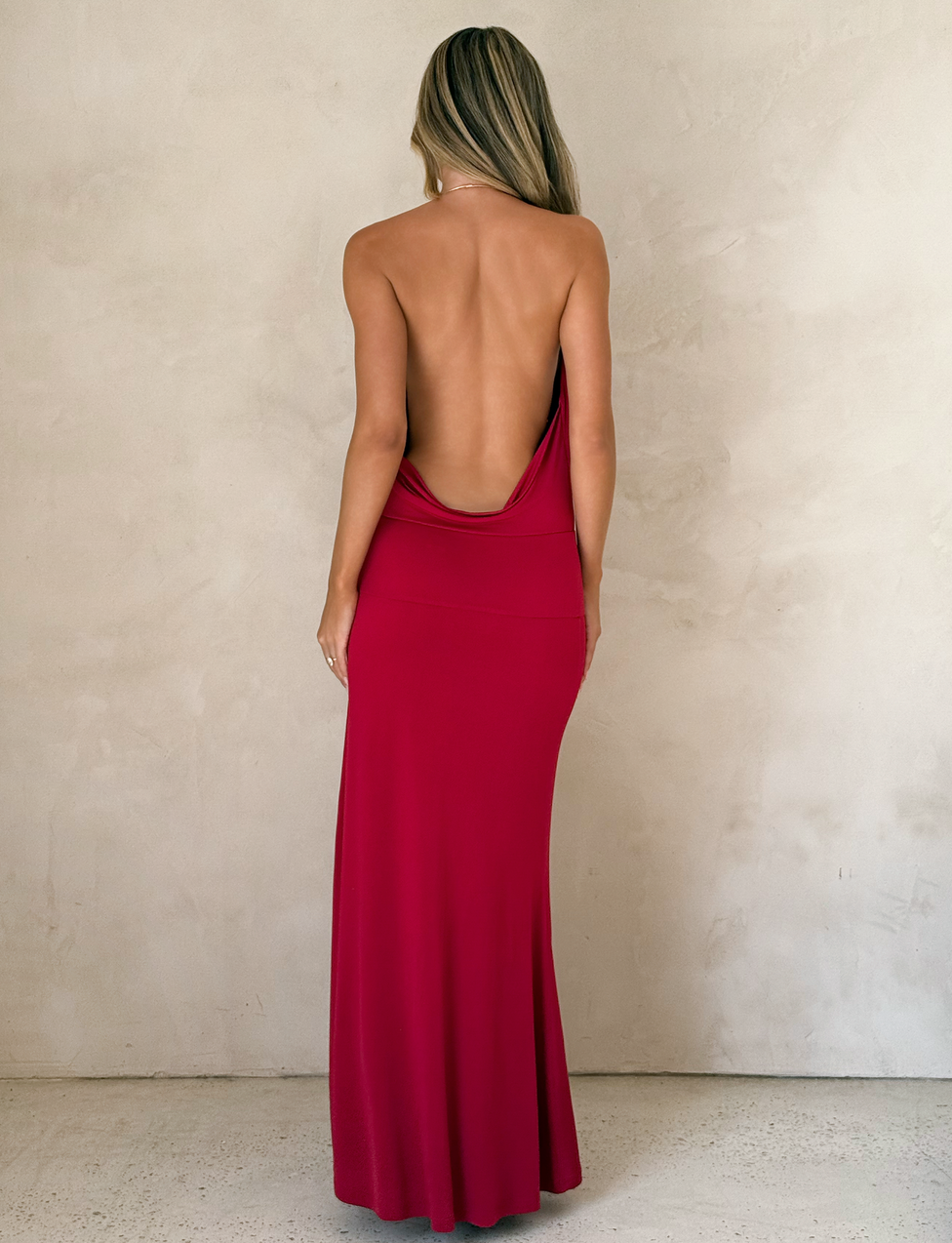 A woman with long blonde hair faces a textured beige wall, wearing the TIGERMIST TARRIN MAXI DRESS in red, featuring a floor-length silhouette, cowl neckline, daring thigh-high slit, and low draped back.