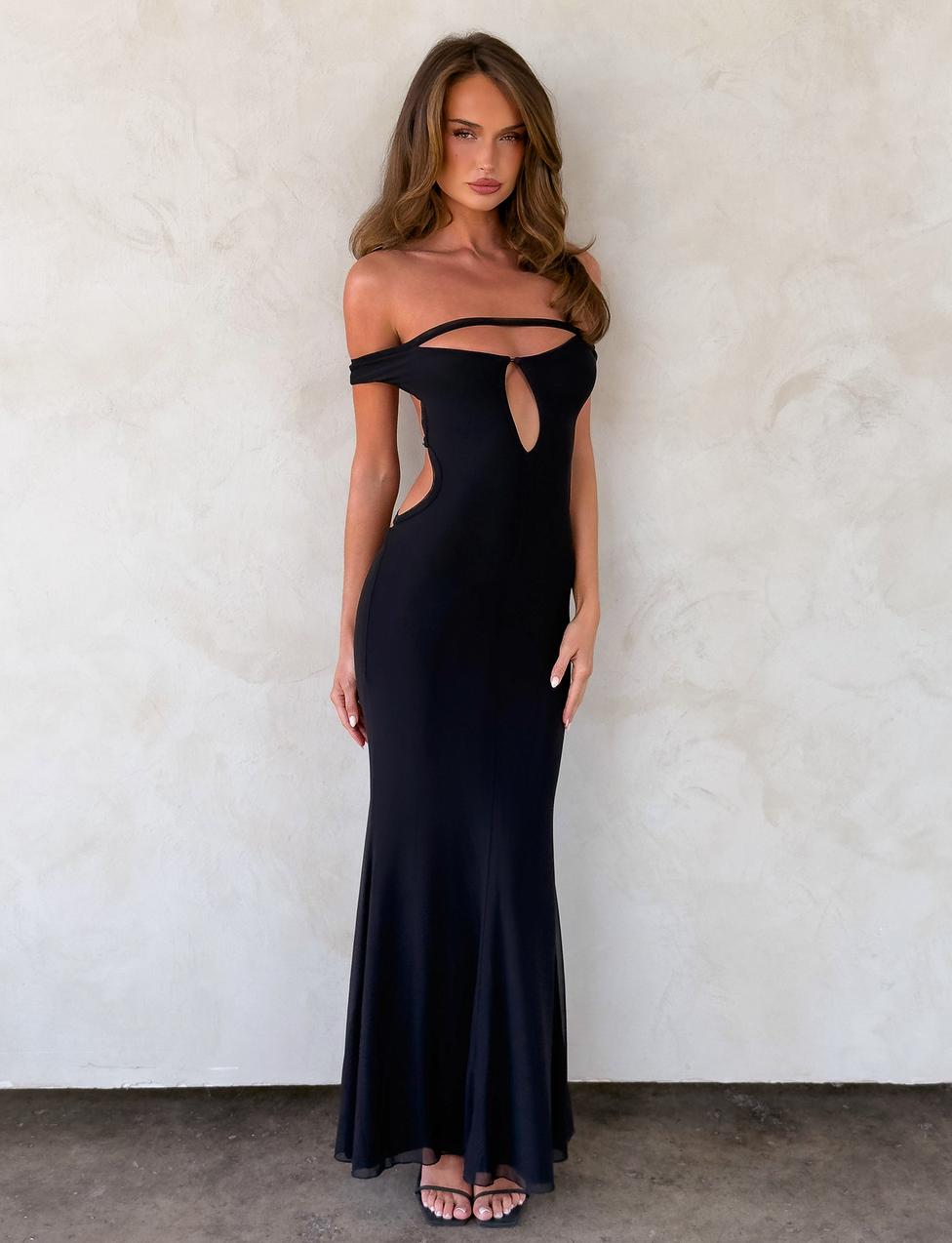 A woman with long, wavy brown hair wears the TIGERMIST KERRIGAN MAXI DRESS in black, featuring off-shoulder and front and side cutout details, paired with strappy sandals against a light textured wall.