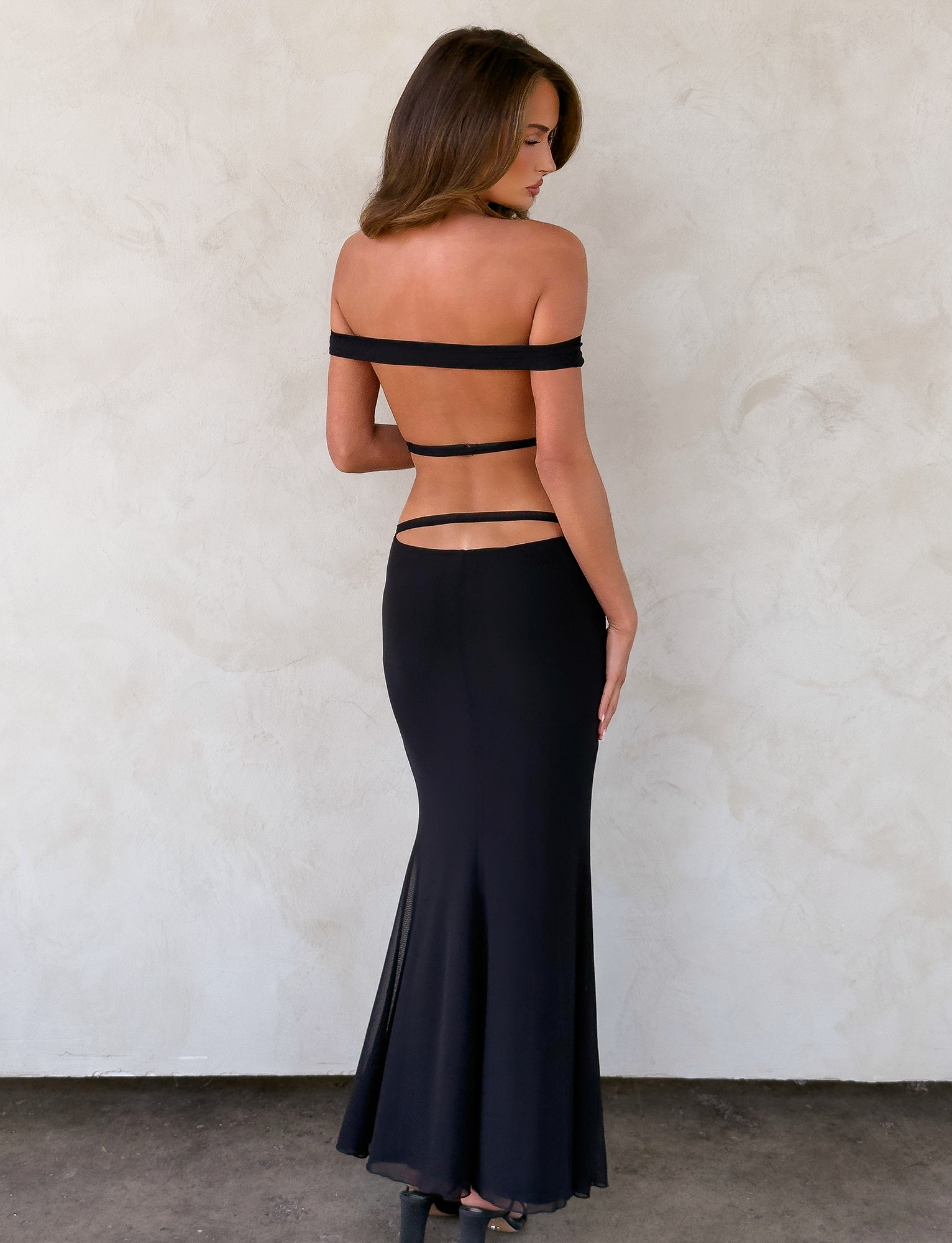 A woman faces away from the camera wearing the TIGERMIST KERRIGAN MAXI DRESS in black, a sleeveless style with a fitted bodice, open back, and thin straps, set against a light textured wall.