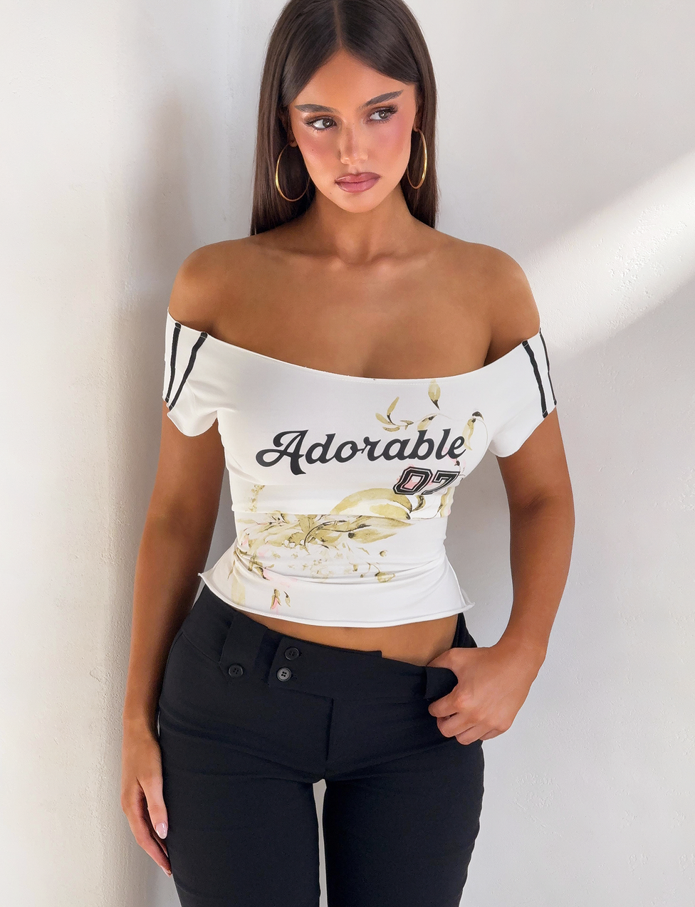 A woman with long brown hair, gold hoop earrings, and black high-waisted pants stands against a white wall, wearing the TIGERMIST FYNN TEE - WHITE, an off-shoulder style with "Adorable 07" and floral print, one hand by her side.