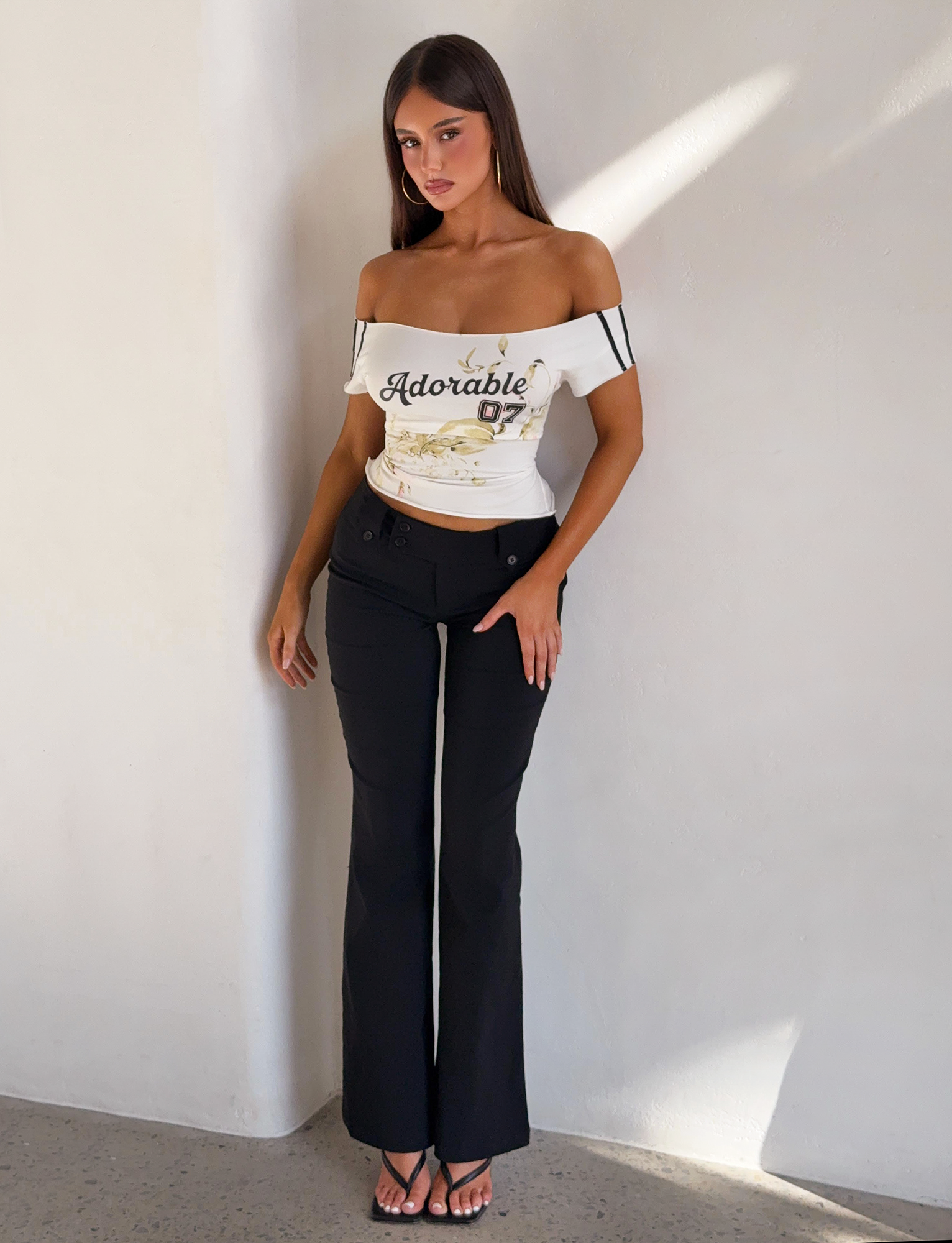 A woman with long brown hair poses indoors against a white wall, wearing the TIGERMIST FYNN TEE in white—an off-shoulder baseball top with "Adorable 07" text—paired with black pants and stylish black heeled sandals.
