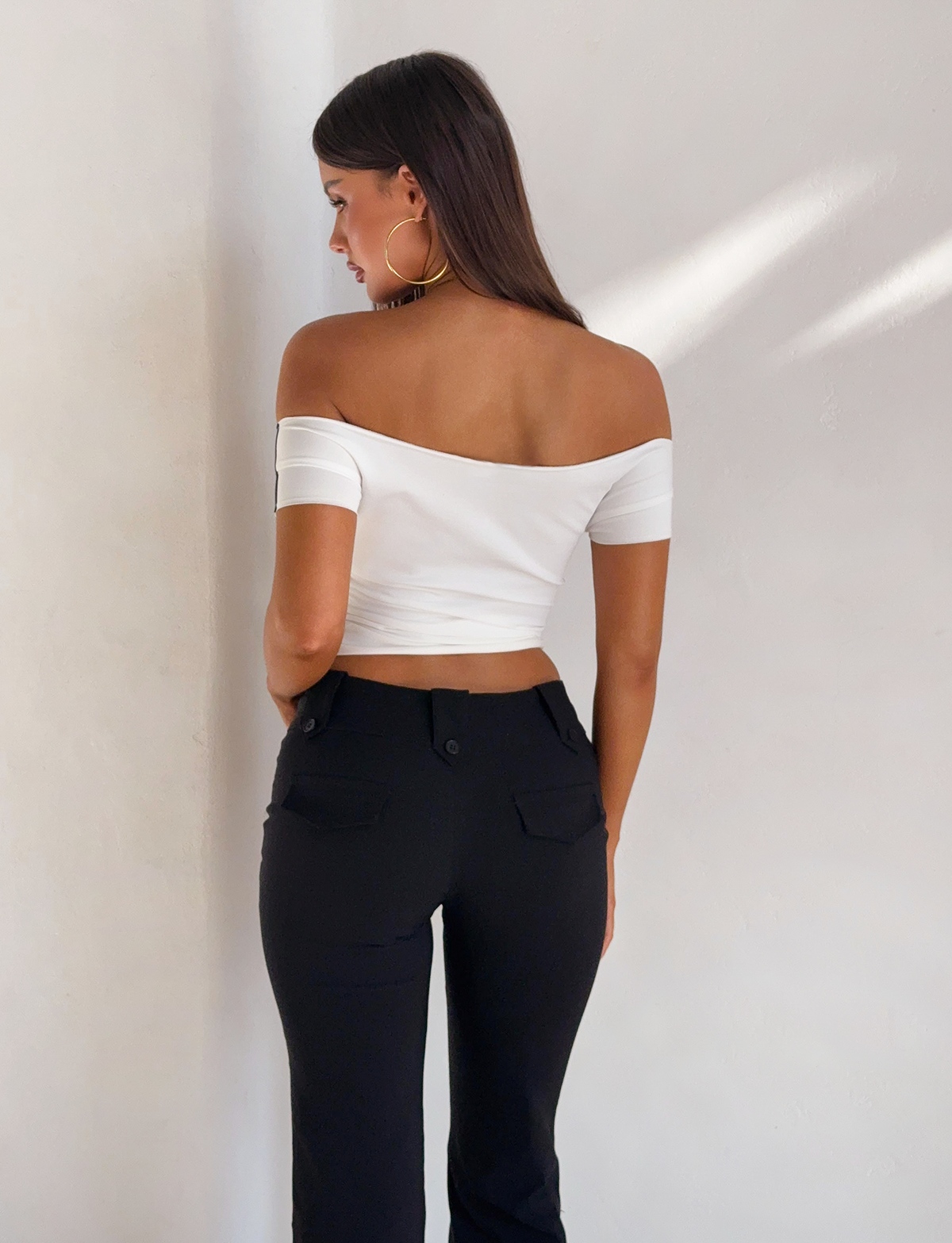 A woman with long brown hair stands facing away, wearing the TIGERMIST FYNN TEE - WHITE off-shoulder top and black high-waisted pants, paired with large hoop earrings, against a plain white wall.