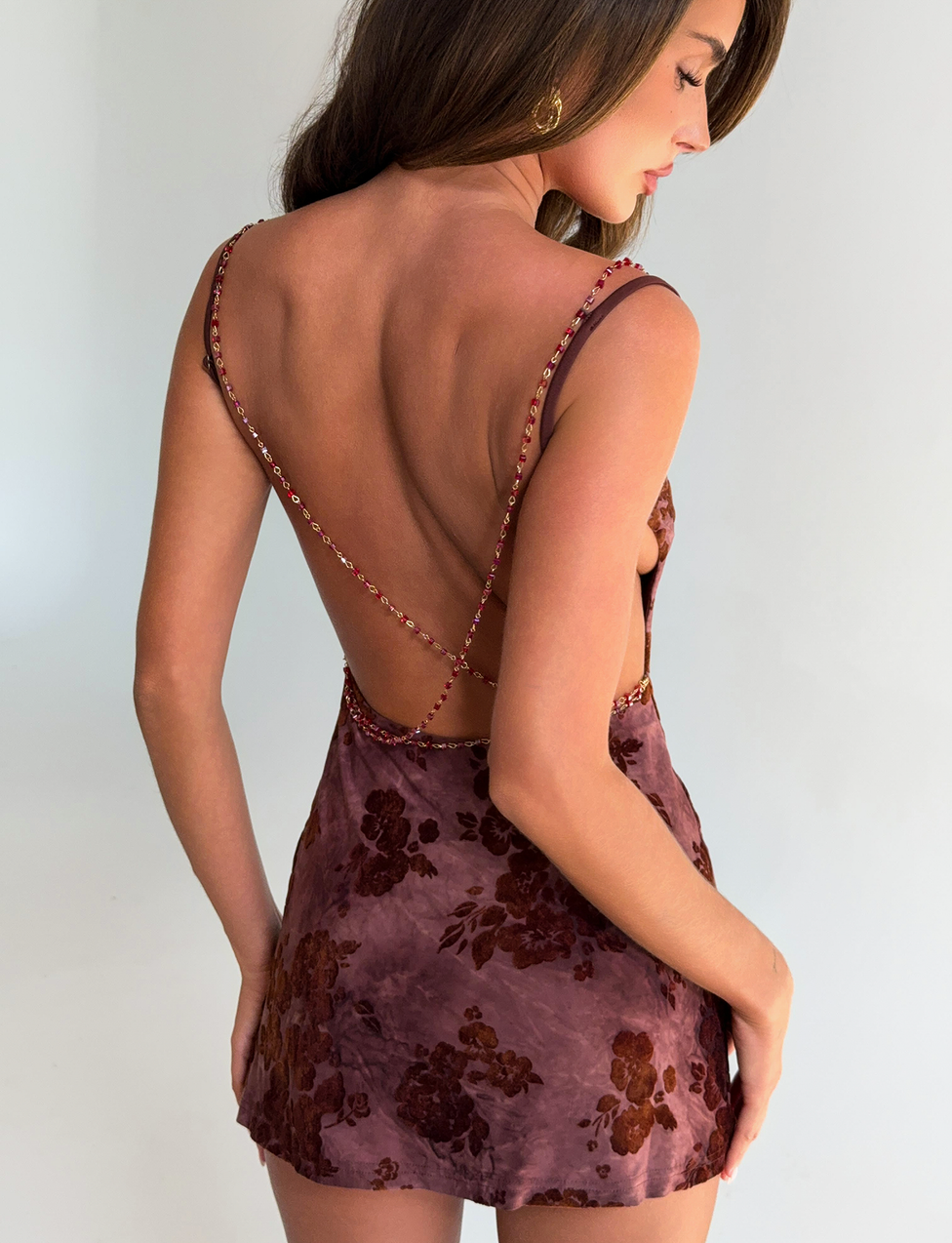 A woman with long brown hair models the TIGERMIST SPARROW MINI DRESS - CHOCOLATE BURNOUT, featuring thin straps and a floral pattern, standing against a plain light background and turned to show her back and side profile.