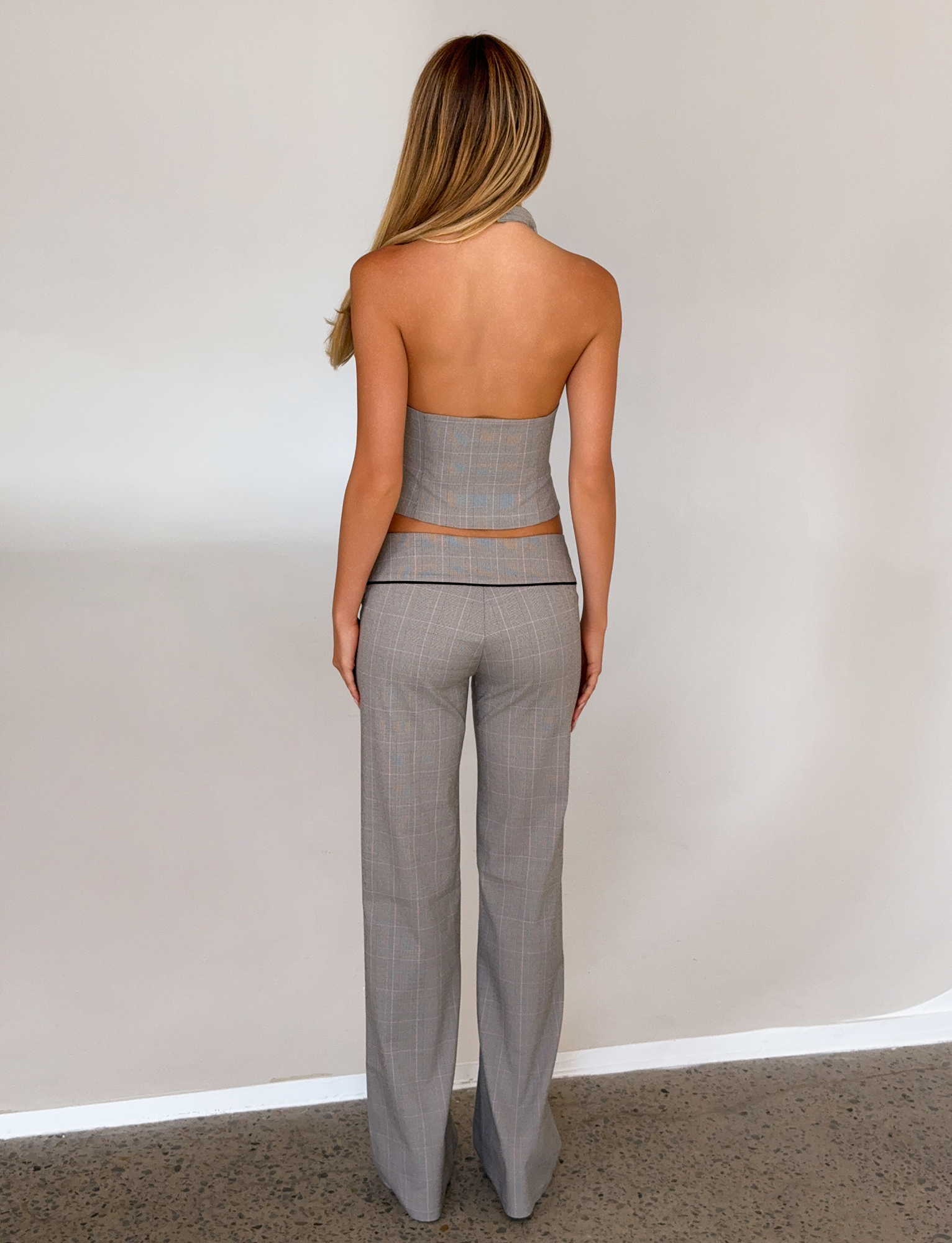 A woman with long blonde hair stands indoors against a neutral wall, wearing TIGERMIST's TERESIA PANT in grey check, paired with a matching backless halter top.