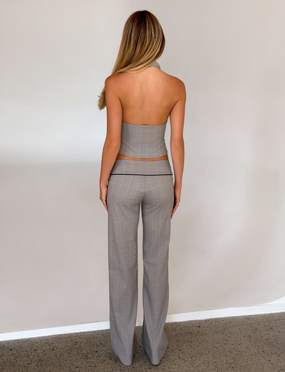 A woman with long blonde hair stands indoors against a neutral wall, wearing TIGERMIST's TERESIA PANT in grey check, paired with a matching backless halter top.