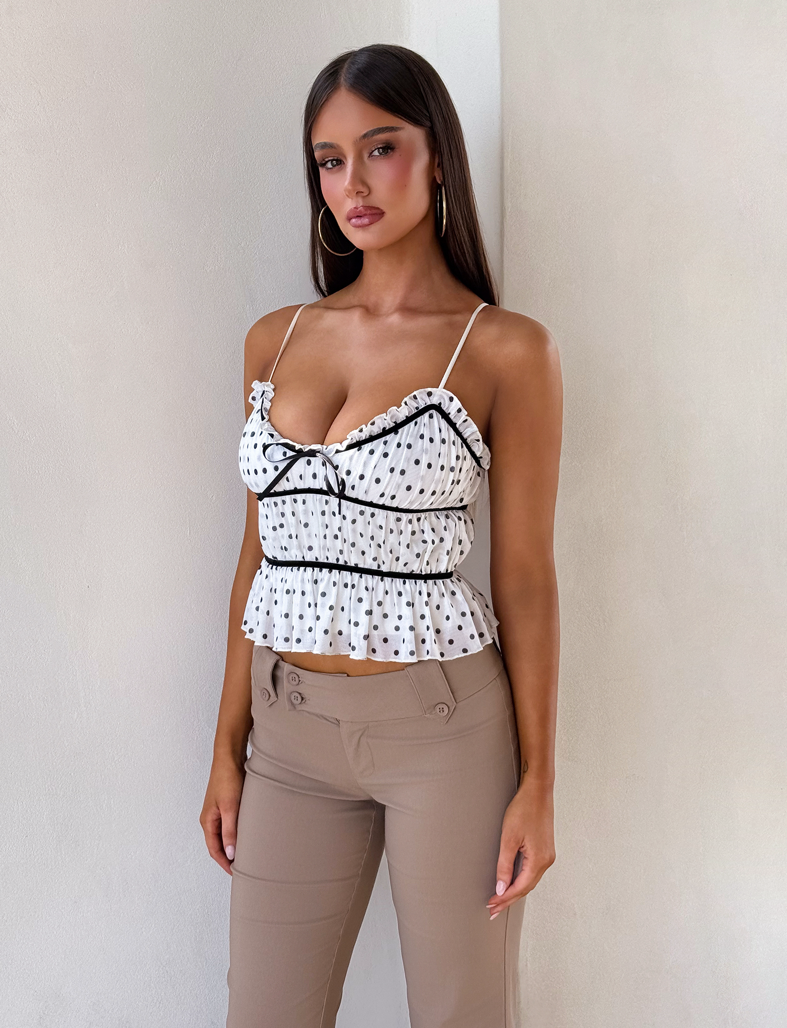 A woman with long brown hair stands against a light wall, wearing the TIGERMIST LIORA TOP - POLKA DOT, a white cami with ruffles and adjustable straps, paired with beige pants. She accessorizes with hoop earrings and has a neutral expression.