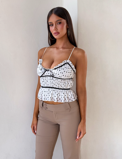 A woman with long brown hair stands against a light wall, wearing the TIGERMIST LIORA TOP - POLKA DOT, a white cami with ruffles and adjustable straps, paired with beige pants. She accessorizes with hoop earrings and has a neutral expression.
