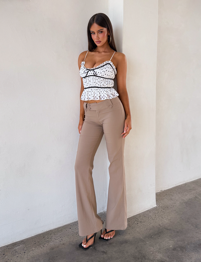 A woman poses against a light wall in the TIGERMIST LIORA TOP - POLKA DOT with adjustable straps and a black bow, styled with beige high-waisted flared pants and black heeled sandals.