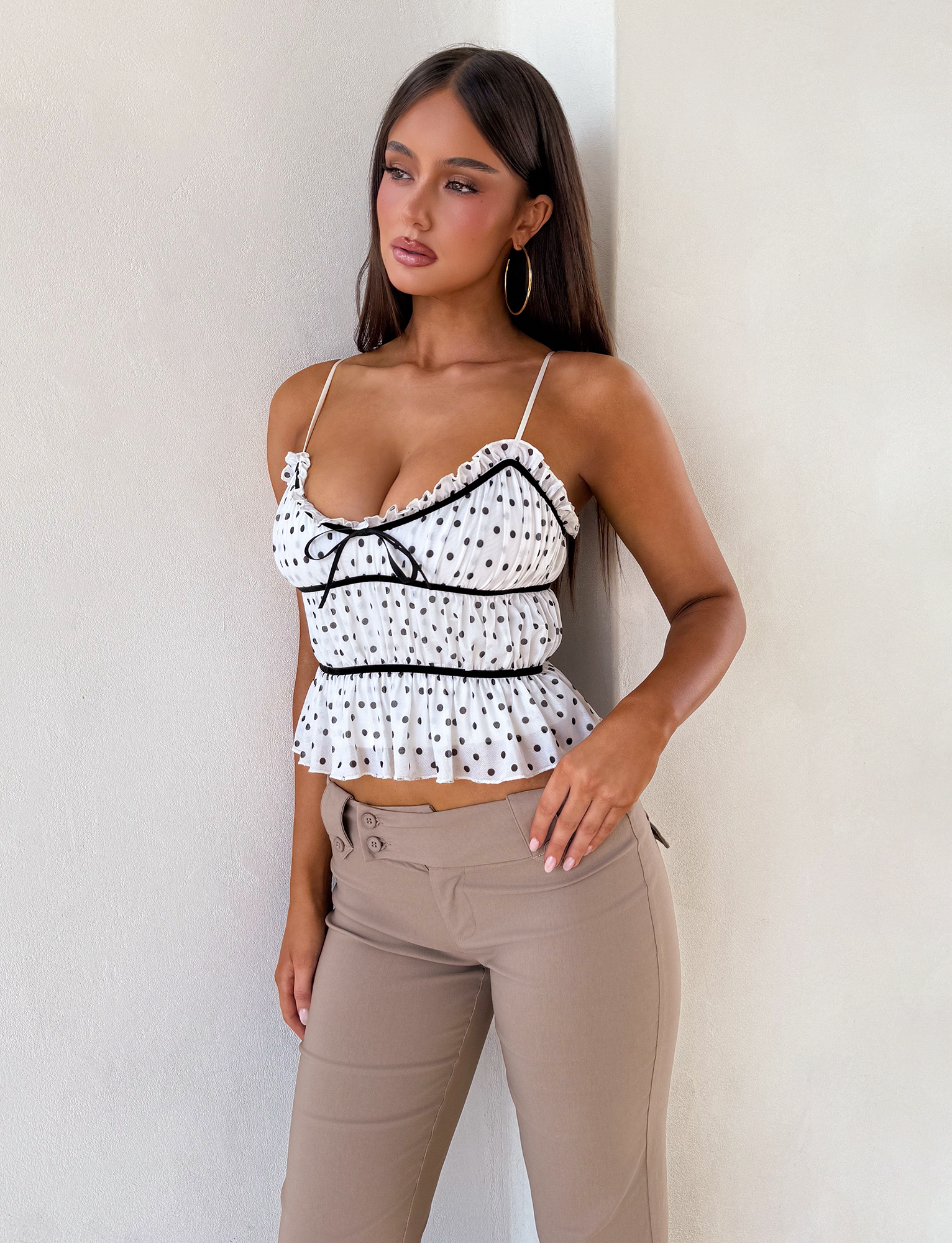A woman with long brown hair wears hoop earrings and the TIGERMIST LIORA TOP - POLKA DOT, a white ruffled cami with adjustable straps and black trim, paired with light beige pants. She stands against an off-white wall, looking to the side.